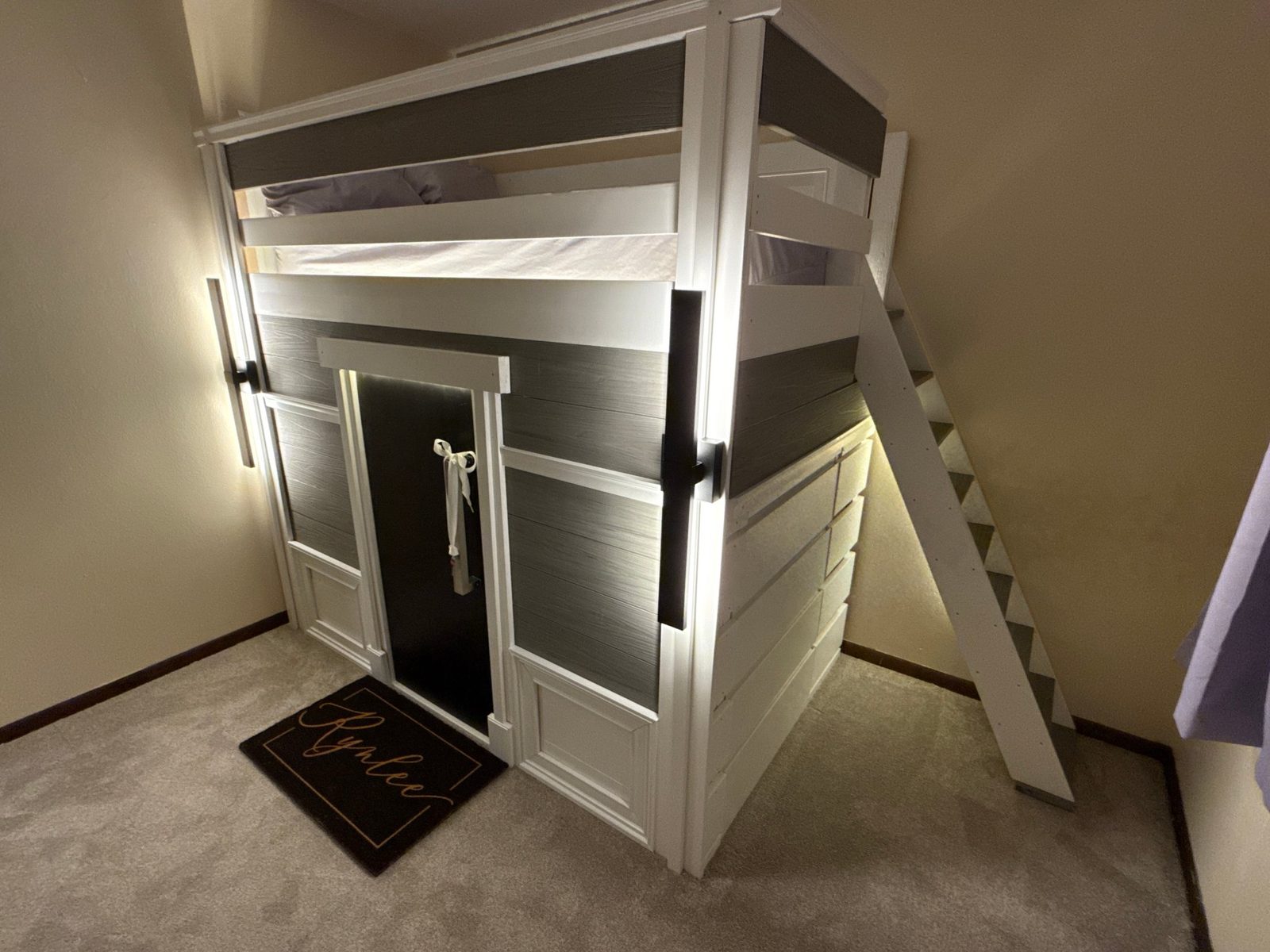 Three-quarter view of the finished playhouse at night — lit entry, black front door, staircase, and storage fitted beneath
